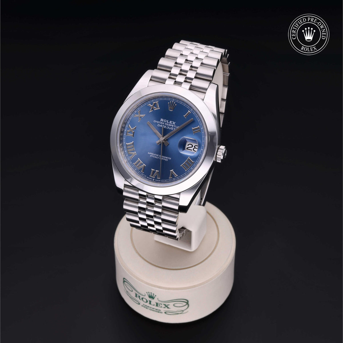 Silver Rolex wristwatch with a blue dial and Roman numeral markers displayed on a branded white stand, featuring a stainless steel Jubilee bracelet and date window.