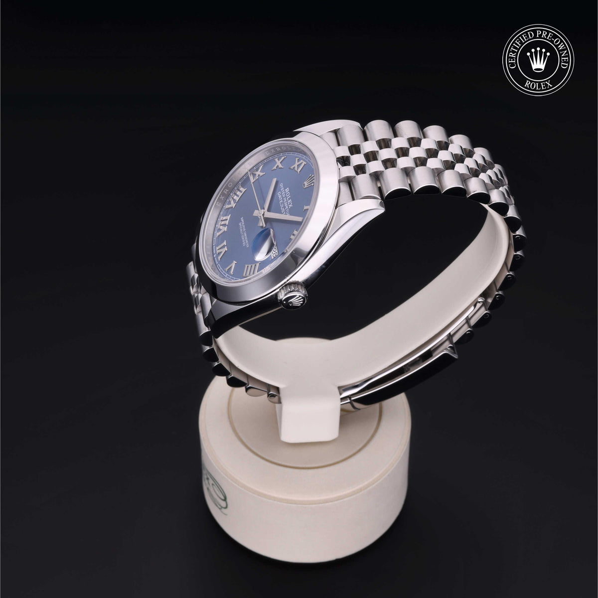 Rolex luxury watch with silver Jubilee bracelet and blue dial featuring Roman numerals, displayed on a white stand with a certified pre-owned Rolex logo against a black background.