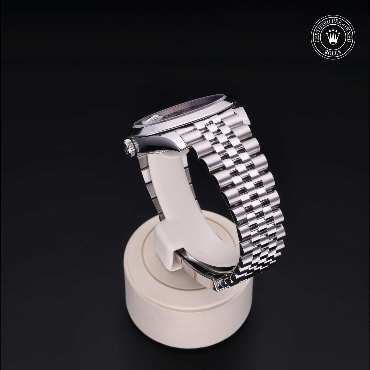 Side view of a Rolex wristwatch with a stainless steel Jubilee bracelet displayed on a white circular stand, accompanied by a certified pre-owned Rolex logo in the corner.