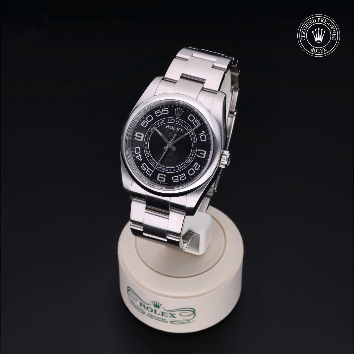 Rolex stainless steel wristwatch with a black dial featuring large white Arabic numerals, displayed on a white branded stand against a black background, certified pre-owned.