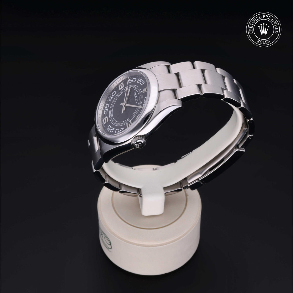 Silver Rolex wristwatch with a black dial and numeric hour markers displayed on a white circular stand against a black background, featuring a Certified Pre-Owned Rolex logo.