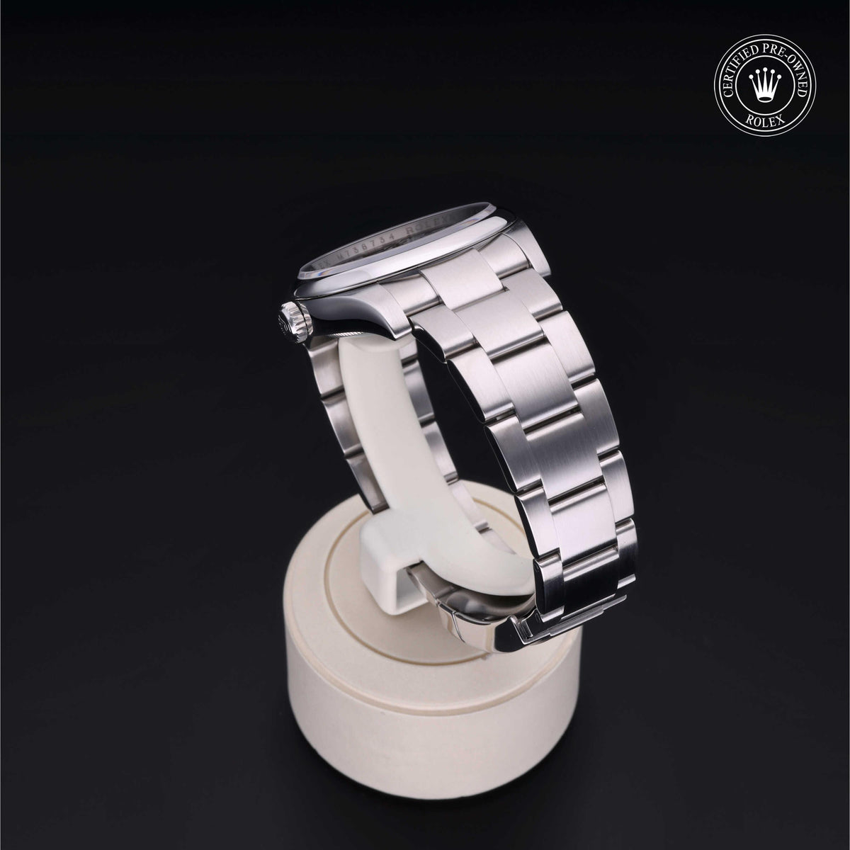 Side view of a polished stainless steel Rolex watch with an Oyster bracelet displayed on a white stand against a dark background, featuring a certified pre-owned emblem.