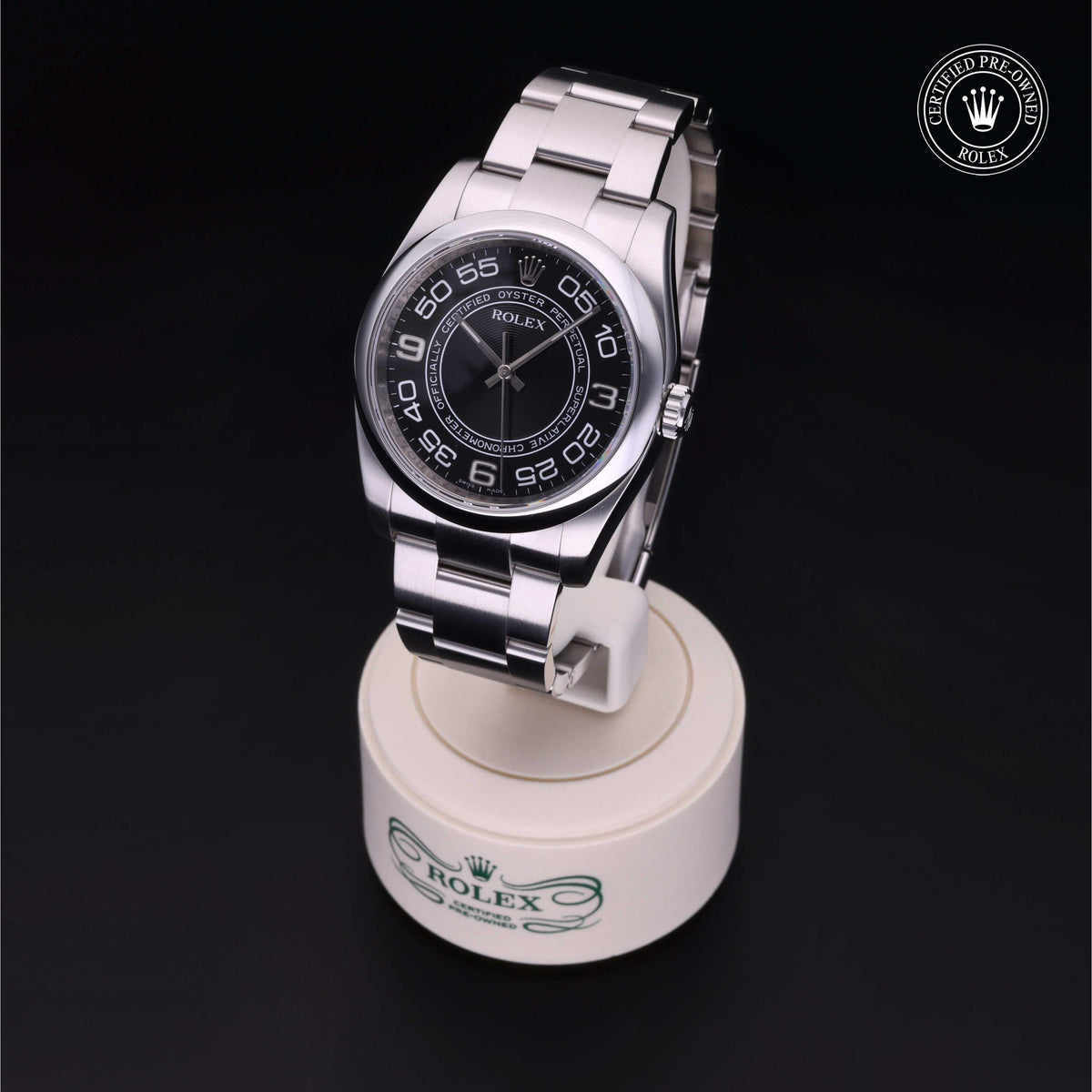 A Rolex stainless steel wristwatch with a black dial and numeric hour markers displayed on a branded white stand against a black background, featuring a certified pre-owned Rolex seal.