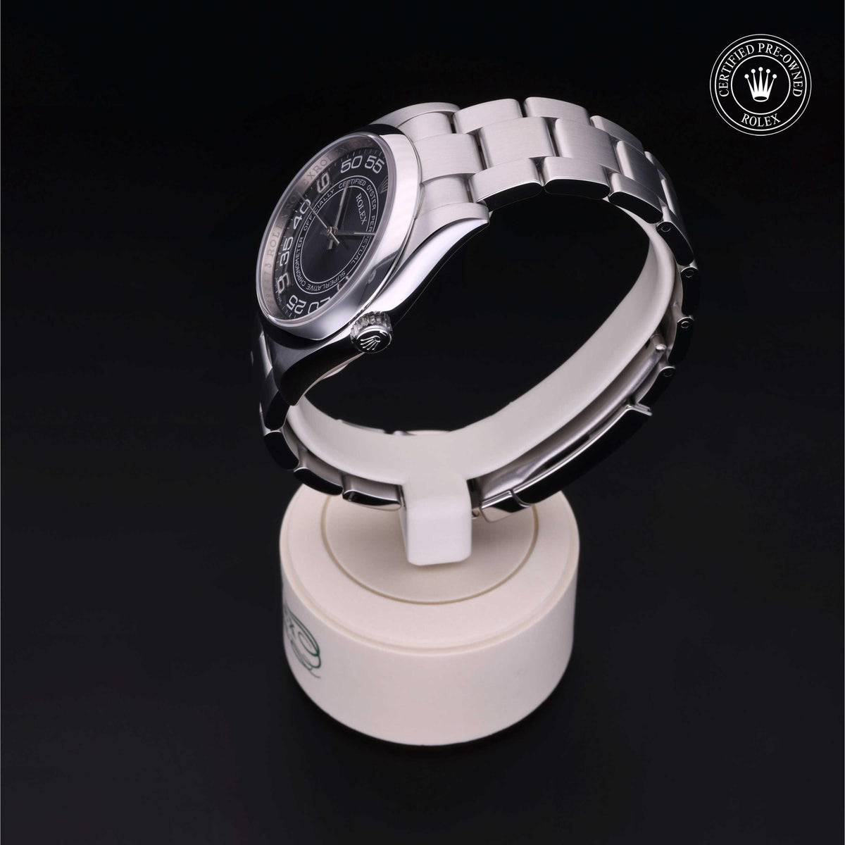 Silver stainless steel wristwatch with a black dial and silver numerals displayed on a white round stand against a dark background, featuring a certified pre-owned Rolex emblem.