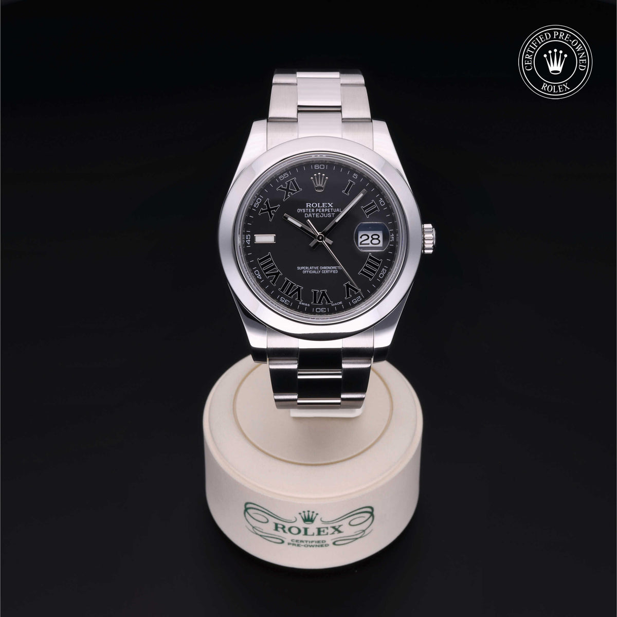 Close-up of a Rolex watch with a black dial featuring Roman numerals and a date window, displayed on a branded stand against a dark background.