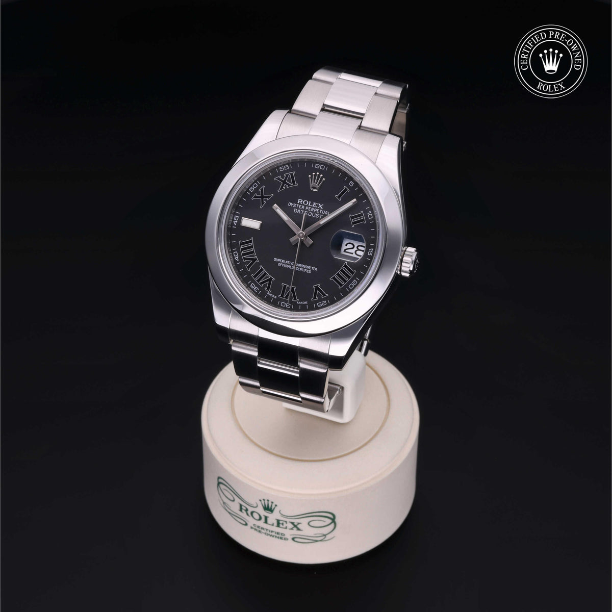 Silver Rolex wristwatch with black dial, Roman numeral hour markers, date display, and stainless steel bracelet resting on a branded watch stand against a black background.