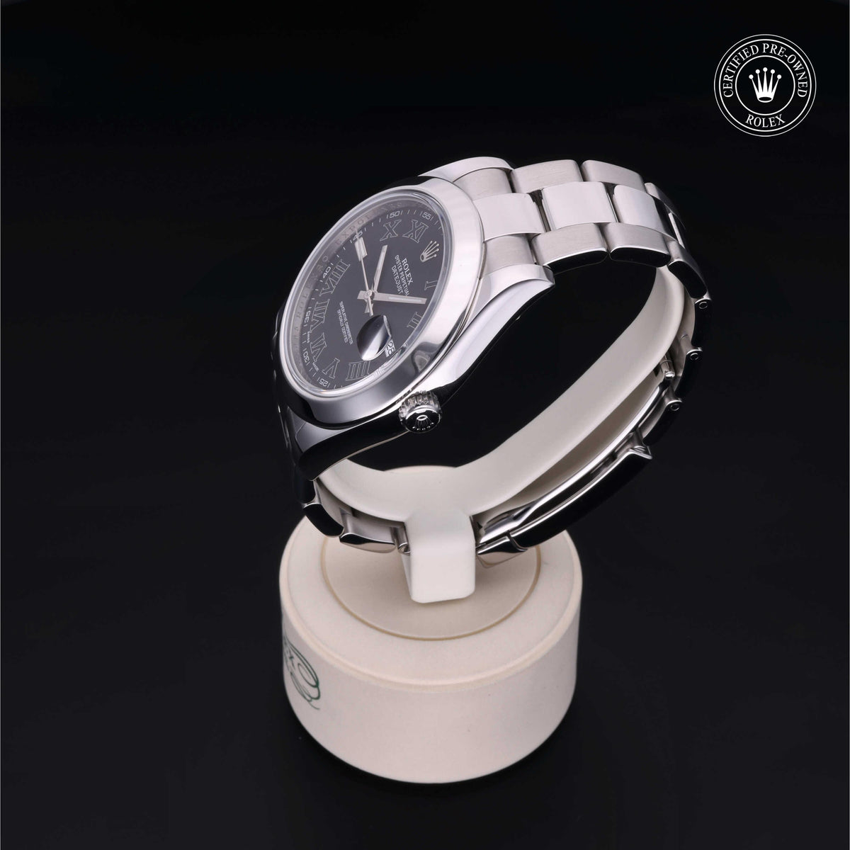 Silver Rolex wristwatch with a black dial and Roman numerals displayed on a white round stand against a black background, marked as Rolex Certified Pre-Owned.